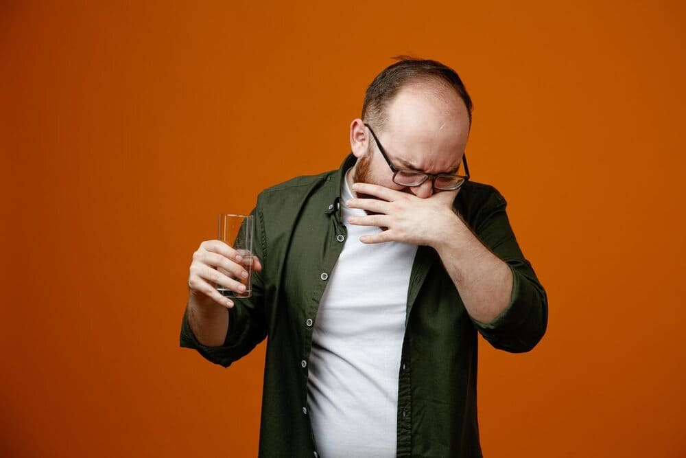 bearded-man-in-casual-clothes-wearing-glasses-holding-a-glass-of-water-looking-confused-and-disappointed-standing-over-orange-background