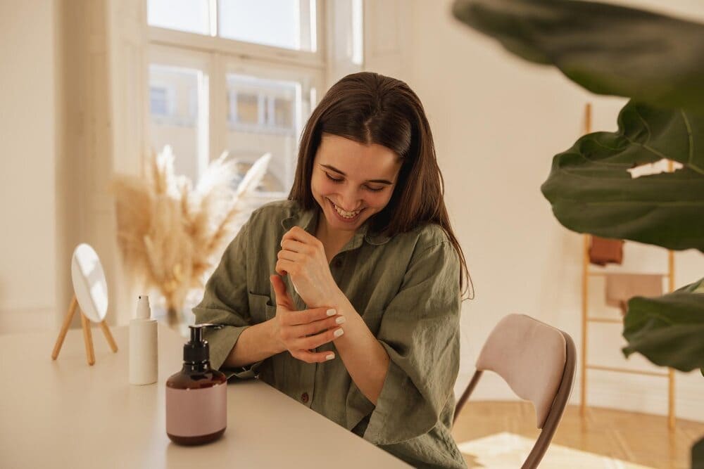 cheerful-young-caucasian-woman-applying-cream-on-her-hands-while-sitting-at-the-table-in-the-light-room-brunette-girl-with-smooth-skin-wears-shirt-wellness-concept