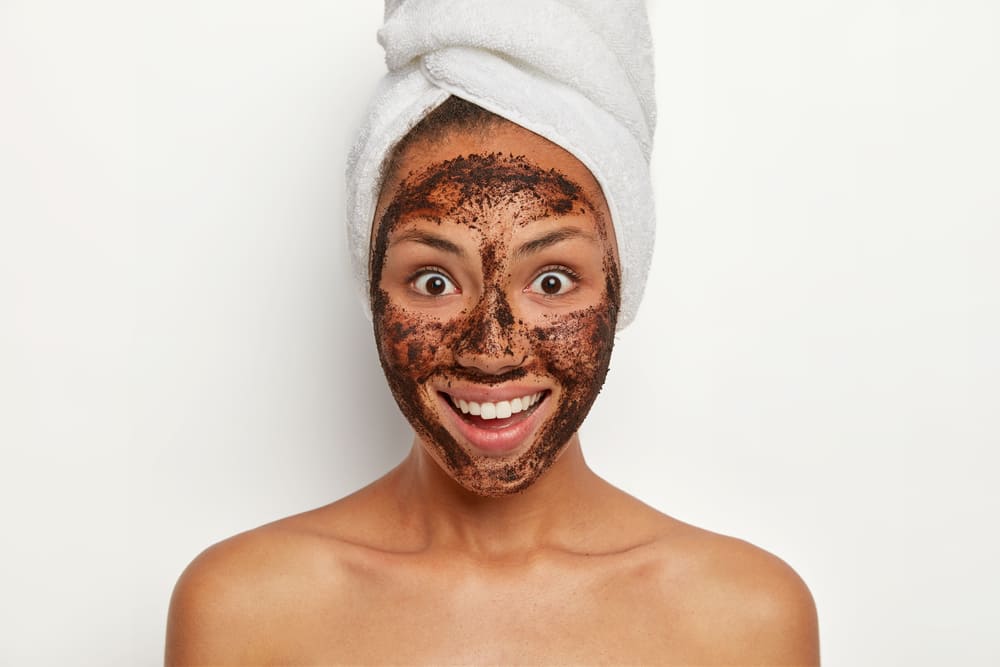 people-skin-care-beauty-concept-smiling-dark-skinned-female-model-cleans-skin-with-coffee-scrub-looks-gladfully-smiles-broadly-has-wrapped-towel-head