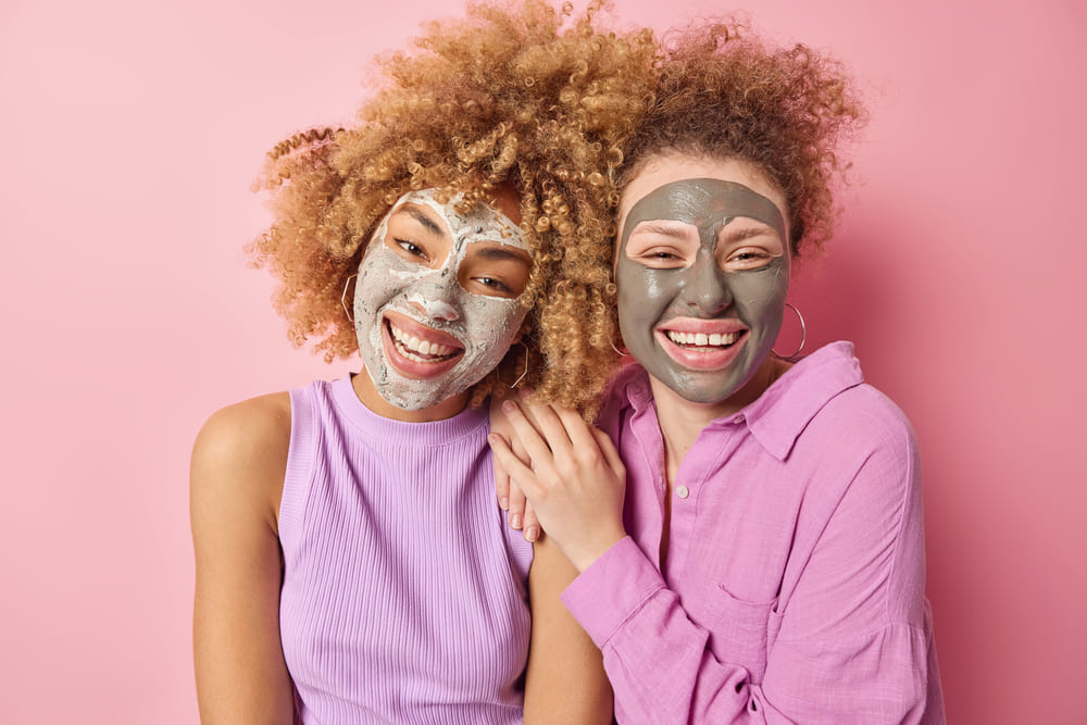positive-women-with-curly-bushy-hair-smile-gladfully-apply-beauty-nourishing-masks-stand-each-other-Women-Happy-to-Know-Benefits-of-Face-Maskswellness