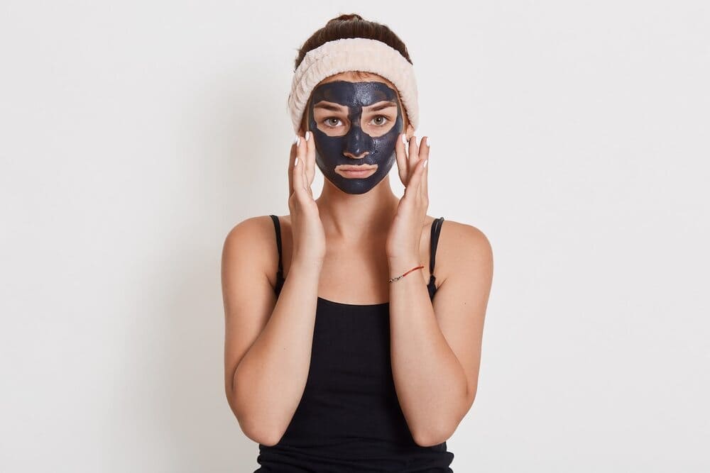 unhappy-young-housewife-with-black-cosmetic-mask-on-her-face-standing-isolated-on-the-white-wall-touching-her-cheeks-with-her-fingers