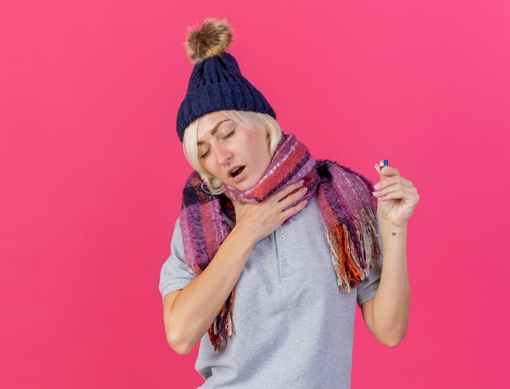 young-blonde-sick-woman-was-dissatisfied-with-hat-and-winter-scarf-puts-her-hand-on-her-neck-and-holds-medical-pills-isolated-on-pink-wall