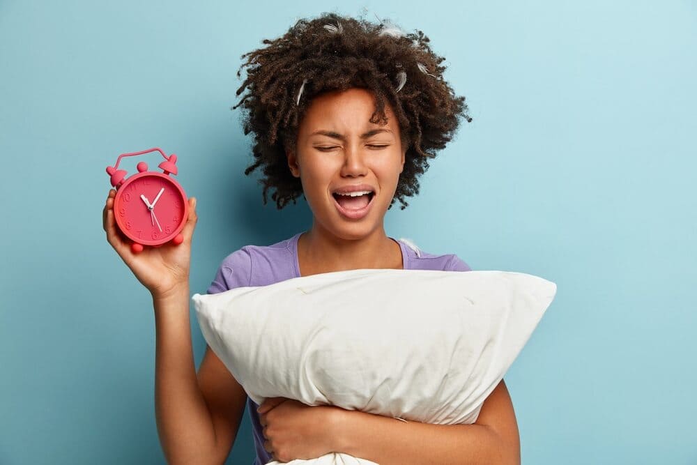 young-brunette-with-feathers-in-her-hair-holding-a-pillow-and-an-alarm-clock