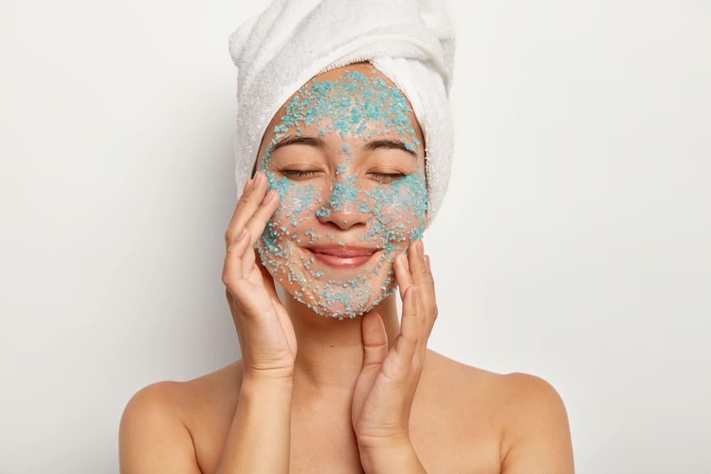 young-cheerful-woman-applies-natural-scrub-face-touches-cheeks-keeps-eyes-closed-wears-towel-has-beauty-procedures-after-glad-to-know-the-Benefits-of-Exfoliating